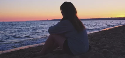 A girl sitting quietly on the beach, looking out at the calm ocean horizon.